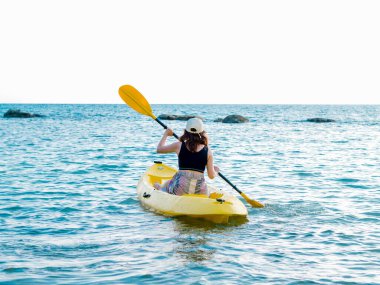 Siyah kolsuz tişörtlü Asyalı bir kadın ve sarı kano kayığında kanoyla denize kürek çekiyor. Mutlu kadın deniz manzaralı arka planda eğlenceli aktiviteler, tatil gezisi.