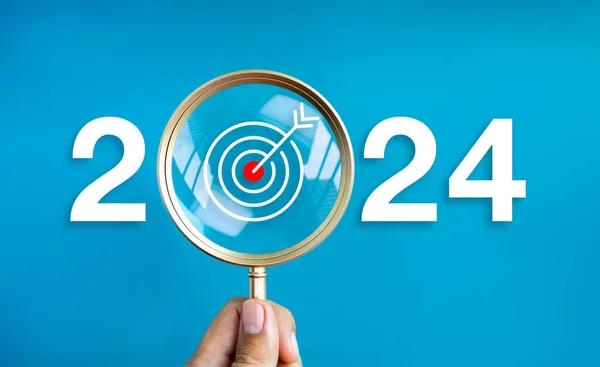 Happy new year 2024 with business concept banner. The big white 2024 year number with target dart icon inside the golden magnifying glass in hand on blue background. Trends, plan and goals concepts.