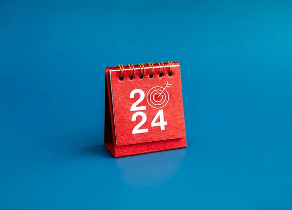 Happy new year 2024 banner background. 2024 numbers year with target dart icon on red small desk calendar cover standing on blue background, minimal style. Business goals plan and success concepts.