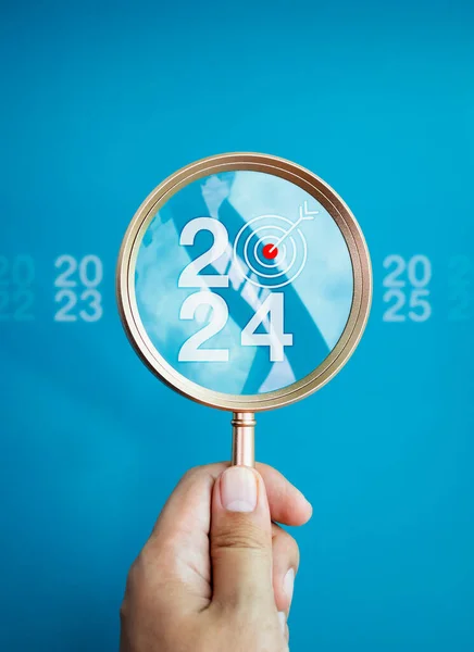 Trends searching, business goals and Happy New Year 2024 concepts. The big white 2024 year number with target icon inside the gold magnifying glass holding by hand on blue background, vertical style.