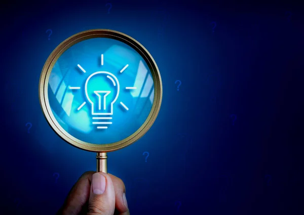 Business solutions, searching content and idea, creative inspiration and find keywords concepts. Lightbulb icon in magnifying glass lens in hand on dark blue background with many question mark symbol.