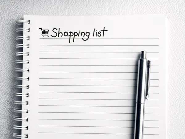 Shopping list, handwriting note text on empty notebook page. Template white paper sheet with line and shopping cart icon on spiral notepad with a silver pen on white leather background.