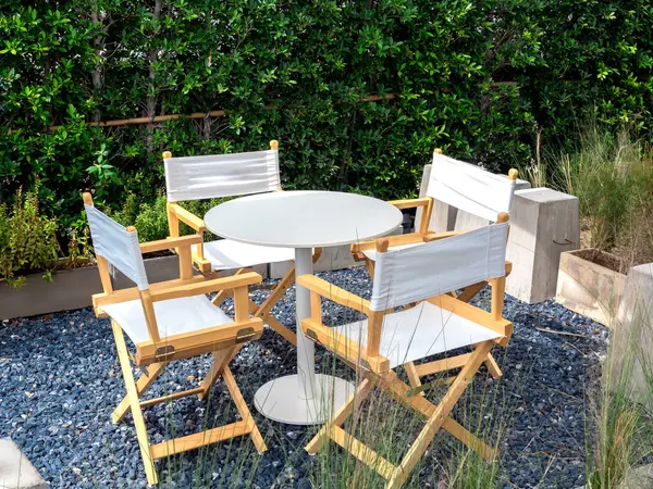 Four empty director chairs, fabric and wood materials with round shape table at outdoor garden on blue gravel stone floor on green tree, bush background. Table set relaxing loft style in the garden.