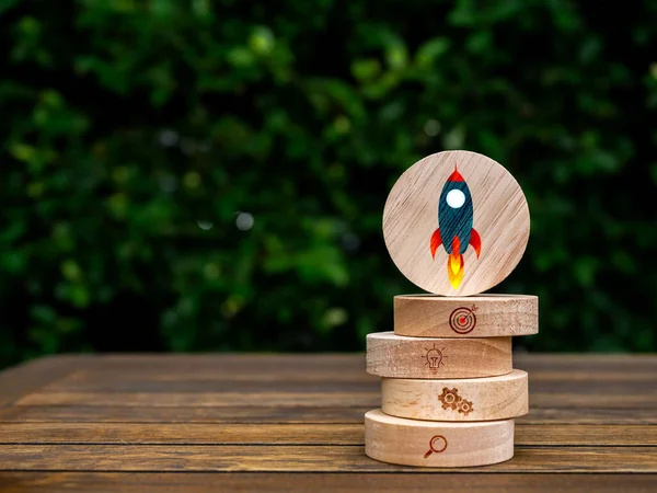Rocket symbol on round wood on top of business strategy icons on wooden block stack on table on green leaves background with copy space. Small business building, startup marketing progress concepts.