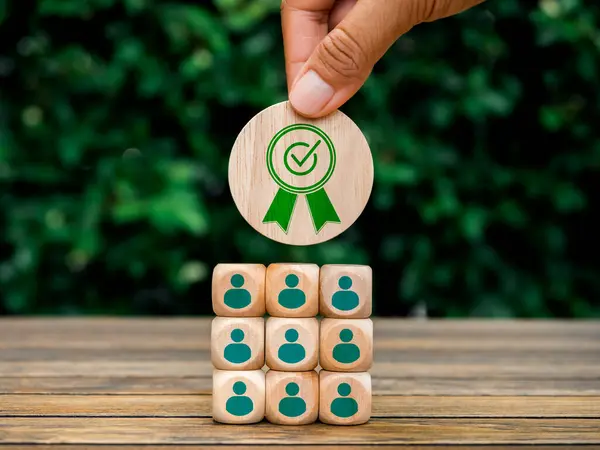 Best quality team and business service success concept. Hand put green warranty icon on wooden cube block on blocks stack with people symbol on wood table and natural green leaves background.