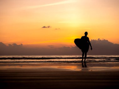Adamın silueti sörf tahtası tutuyor, karanlık deniz kıyısında fotokopi alanı ile yürüyor. Günbatımı gökyüzünde duran sörfçü deniz suyu arka planında. Akşam vakti yaz kumsalı. Alacakaranlık deniz manzarası.