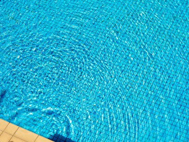 Yüzme havuzu arka planındaki mavi mozaik fayanslar üzerinde berrak suda dalgalanan bulanık yuvarlak görünüm. Güneşli bir günde havuz kenarındaki boş alan, yaz tatili..