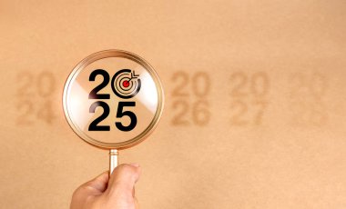 İş konsepti afişiyle 2025 yılınız kutlu olsun. Açık kahverengi arka planda büyüteç mercekli hedef dart ikonu olan büyük siyah 2025 yıllık sayı. İş eğilimi, çözüm planı konsepti.