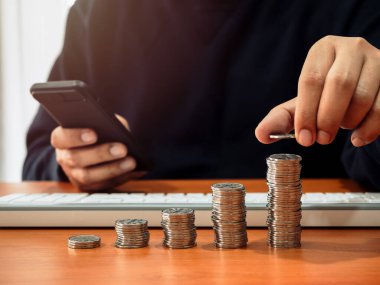 Çubuk grafik çizelgesi olarak istiflenmiş bozuk paralar ahşap masada cep telefonu tutan kişi tarafından düzenlenir. Yatırım, kar geliri, bağış, iş büyümesi, açık tasarruf hesabı online kavramları.