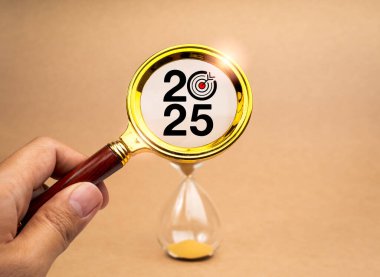 2025 Mutlu yıllar. Yükleme, iş ve teknoloji trendleri ve başarı hedefleri konsepti. 2025 Numara ve hedef simgesi elinde altın büyüteçle, kahverengi arkaplanda kum saatine yakınlaşırken.