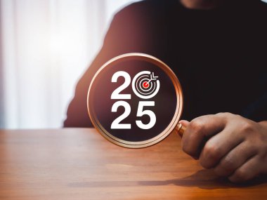 İş konsepti afişiyle 2025 yılınız kutlu olsun. Büyüteç merceğinde hedef simgesi olan 2025 yıllık rakamlar. Masanın üzerinde kişinin elinden tutulmuş, koyu arkaplan. Hedef ve başarı kavramları için planlama.