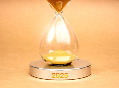 Yakında 2025 yılına geliyoruz, iş ve teknoloji kavramı. Kapalı kum saati, içinde altın kumu olan yuvarlak gümüş krom podyum, kahverengi arka planda 2025 yıllık takvim numarası, minimalist..