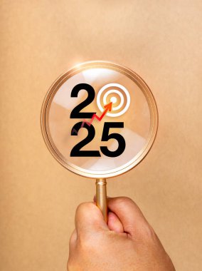İş amaçlı trend konseptiyle 2025 yılınız kutlu olsun. Kırmızı okla vurulan hedef simgeye sahip 2025 takvim numarası, dikey kahverengi zemin üzerinde elle tutulan altın büyüteç içinde odaklanmış..
