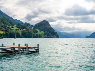 Brienz Gölü 'nde kimsenin olmadığı boş ahşap iskele manzarası. Yeşil tepedeki köy, dağlar, bulutlu arka planda su. İsviçre 'nin Iseltwald şehrinde ünlü bir yer..