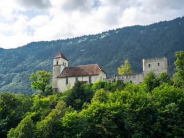 Ringgenberg şatosu kilisesi, Brienz Gölü yakınlarındaki yeşil ağaçlar tepesindeki eski geleneksel binalar, ve dağ ve bulutlu yaz gökyüzü arka planı, deniz yolculuğu manzarası, İsviçre.