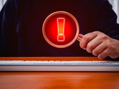 Uyarı, dolandırıcılık, kırmızı ünlem işareti simgesi, sistem hacklenmiş hata işareti, kötü amaçlı yazılım, büyüteç merceğiyle gösterilen dikkat tehlikesi uyarısı, bilgisayarla çalışan kişinin elinde..