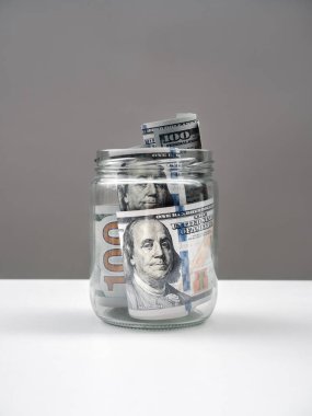 100 dolar nakit para. Benjamin Franklin 'in yüzü cam kavanozda. Beyaz masa ve gri arka planda, dikey şekilde izole edilmiş. Tasarruf, bütçe, vergi, mali konsept.