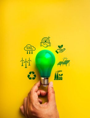 Tasarruf enerjisi, karbon azaltımı, sürdürülebilirlik konsepti. İnsan eli tarafından tutulan yeşil ampul, yenilenebilir işaretler ve koruma sembolleri ile çevrili, sarı arka plan, minimalist.