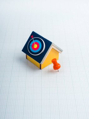 Ev ve arazi konumu, mülk değeri, emlak alım ve satım pazarı ve yatırım kavramları. Grafik kağıdında turuncu raptiye olan minyatür ev modelinin 3D hedef simgesi, dikey arkaplan.