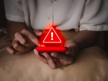 Hacker saldırı algılama alarmı, SMS dolandırıcılığı ve dolandırıcılık aramaları, sahte telefon görüşmeleri konsepti. Kırmızı üçgen uyarı alarmı yaşlı ellerde, karanlık odada akıllı cep telefonunda görünüyor..