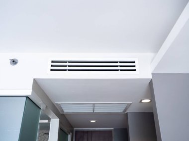 Beyaz otel odasının arka planındaki giriş kapısının üstündeki dar tavana monte edilmiş havalandırma sisteminin ön görüntüsü. Otel odasının havalandırma ızgarası duvarda..