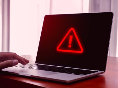Virüs alarmı. Kırmızı üçgen, sistem hacklenmiş hata işareti, kötü amaçlı yazılım, dikkat tehlike sembolü, sistem hatası, masadaki bilgisayar ekranında işadamı eli, kırmızı tonlu uyarı.