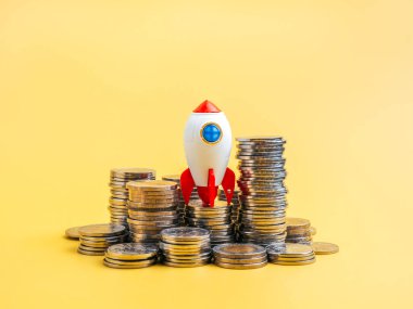 İş büyümesi ve başarı, girişim işi, finansal planlama, bankacılık ve finans, yatırım konsepti. Başlangıç roketi oyuncağı, sarı arka planda izole edilmiş bir sürü para madeni parasına yerleştirildi.