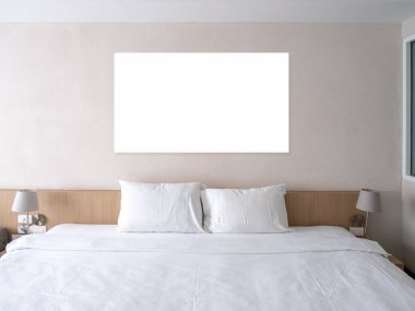 Boş kare çerçeve beyaz duvarda asılı, yastıklı ve temiz battaniyeli tahta yatak, otel odasının arka planında lambalı süsleme, insan yok..