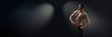 Kaslı bir adam karanlık bir arkaplanda loş ışıkta poz veriyor, geniş bir kopya alanı var. Fitness reklamları ve motivasyon materyalleri için ideal. Geniş pankart.