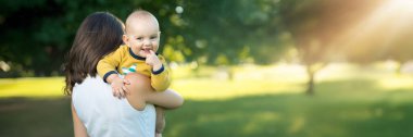 Güneşli bir park ortamında annesinin omzunda neşeli bir bebek. Parlak renkler ve doğal ortam, aile veya ebeveynlik teması için uygun. Kopyalama alanı olan geniş pankart.