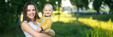 Neşeli bir anne, gülümseyen bebeğini güneşli bir parkta tutuyor. Canlı yeşiller. Aile, ebeveynlik ve açık hava temaları için mükemmel. Kopyalama alanı olan geniş pankart.