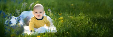 Canlı yeşil bir çayırda, çiçekli bir battaniyenin üzerinde gülümseyen bebek. Parlak renkler ve doğal ışık neşeli bir atmosfer yaratır. Ebeveynlik, yaşam tarzı ve bahar temaları için uygun.