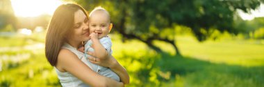 Güneşli bir parkta gülümseyen bebeği tutan bir anne. Sıcak renkler, samimi bir tarz. Aile ve açık hava reklamları için ideal. Kopyalama alanı olan geniş pankart.