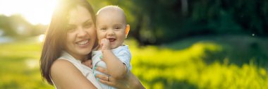 Güneşli bir alanda kucaklayan ve gülümseyen neşeli bir anne ve bebek. Sıcak renkler ve açık hava ortamları aile ve mutluluk temalarını çağrıştırıyor. Kopyalama alanı olan geniş pankart.