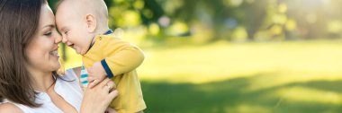 Güneşli bir parkta bebeğini kucaklayan bir anne, içten bir gülümsemeyle. Parlak renkler ve bulanık arkaplan huzurlu bir his yaratır. Aile, ebeveynlik ve yaşam tarzı içeriği için ideal.