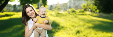 Güneşli bir parkta gülümseyen bebeğini tutan neşeli bir anne. Doğal yeşil tonlara ve davetkar bir atmosfere sahip. Aile ya da yaşam tarzı teması için ideal. Kopyalama alanı olan geniş pankart.