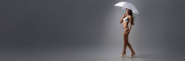 Beyaz bikinili bir şemsiyeyle gri bir arkaplanda poz veren bir model. Zarif ve şık, moda ya da yaz promosyonları için ideal. Kopyalama alanı olan geniş pankart.