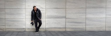 Taş bir duvara yaslanmış siyah kıyafetli şık bir adam. Tarafsız ses tonları, modern görünüş. Moda reklamları ve yazı işleri için ideal. Kopyalama alanı olan geniş pankart.