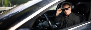 Siyah takım elbiseli adam arabada güneş gözlüğü takıyor. Şık, modern görünüş, şık tasarım, pazarlama, iş dünyası veya otomotiv temaları için uygun. Kopyalama alanı olan geniş pankart.
