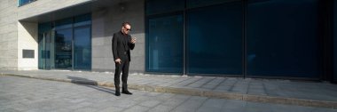 Siyah takım elbiseli şık bir işadamı, akıllı telefon kullanıyor, açık havada. Modern mimari, doğal ışık. İş promosyonları için mükemmel. Kopyalama alanı olan geniş pankart.