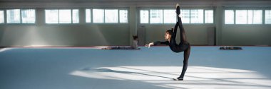 Siyah kıyafetli genç bir jimnastikçi, spor reklamcılığı için mükemmel olan geniş bir spor salonunda bacak esnetme hareketi yapıyor. Kopyalama alanı olan geniş pankart.