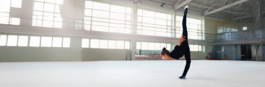 Genç jimnastikçi parlak bir spor salonunda ikiye bölünmüş bir performans sergiliyor. Hafif tonlu modern tarz. Spor, spor ve esneklik promosyonları için ideal. Kopyalama alanı olan geniş pankart.