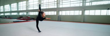 Genç bir jimnastikçi geniş bir spor salonunda bölünme gerçekleştiriyor. Doğal ışık, siyah giysi ve sakin ifade onu spor ve spor reklamları için ideal kılıyor..