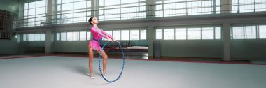 Pembe elbiseli bir jimnastikçi geniş bir spor salonunda çemberle gösteri yapıyor. Parlak renkler ve dinamik duruş. Spor reklamları ve fitness promosyonları için ideal. Geniş pankart.
