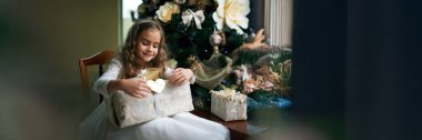 Genç bir kız Noel hediyesini süslü bir ağacın yanında sevinçle açıyor. Konforlu tatil sahnesinde sıcak renkler, şenlikli promosyonlar veya reklamlar için mükemmel. Geniş pankart.