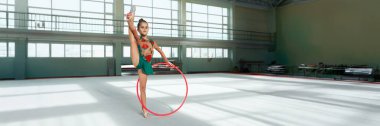 Genç bir jimnastikçi geniş bir spor salonunda çemberle gösteri yapıyor. Görüntü, spor promosyonları için ideal, parlak ışıklandırma, canlı kostüm ve fotokopi alanı içeriyor..