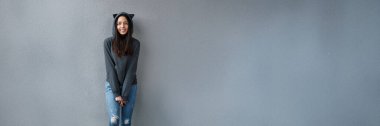 Kedi kulaklı kapüşonlu mutlu genç kadın gri bir duvara gülümsüyor. Minimalist stil ve havalı tonlar geniş bir kopya alanı sağlıyor. Reklam için ideal..
