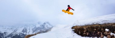 Kırmızı ve sarı teçhizatlı snowboardcu karlı tepelere doğru atlıyor. Canlı renklerle dinamik aksiyon çekimi. Kış sporları promosyonları için mükemmel. Geniş pankart.