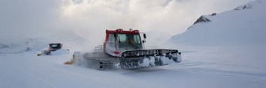 Kızıl bir kar aracı dağlık bir arazide karlı bir pisti düzleştiriyor. Geniş pankart stili, kış sporu reklamları için ideal olan geniş karlı alanlarda fotokopi alanı sergiliyor.
