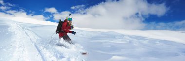 Bir snowboardcu açık mavi gökyüzü ile aydınlık bir günde karlı bir yamaçtan iner. Görüntü canlı bir renk paletine ve geniş bir kopyalama alanına sahiptir. Kış sporları promosyonları için mükemmel..
